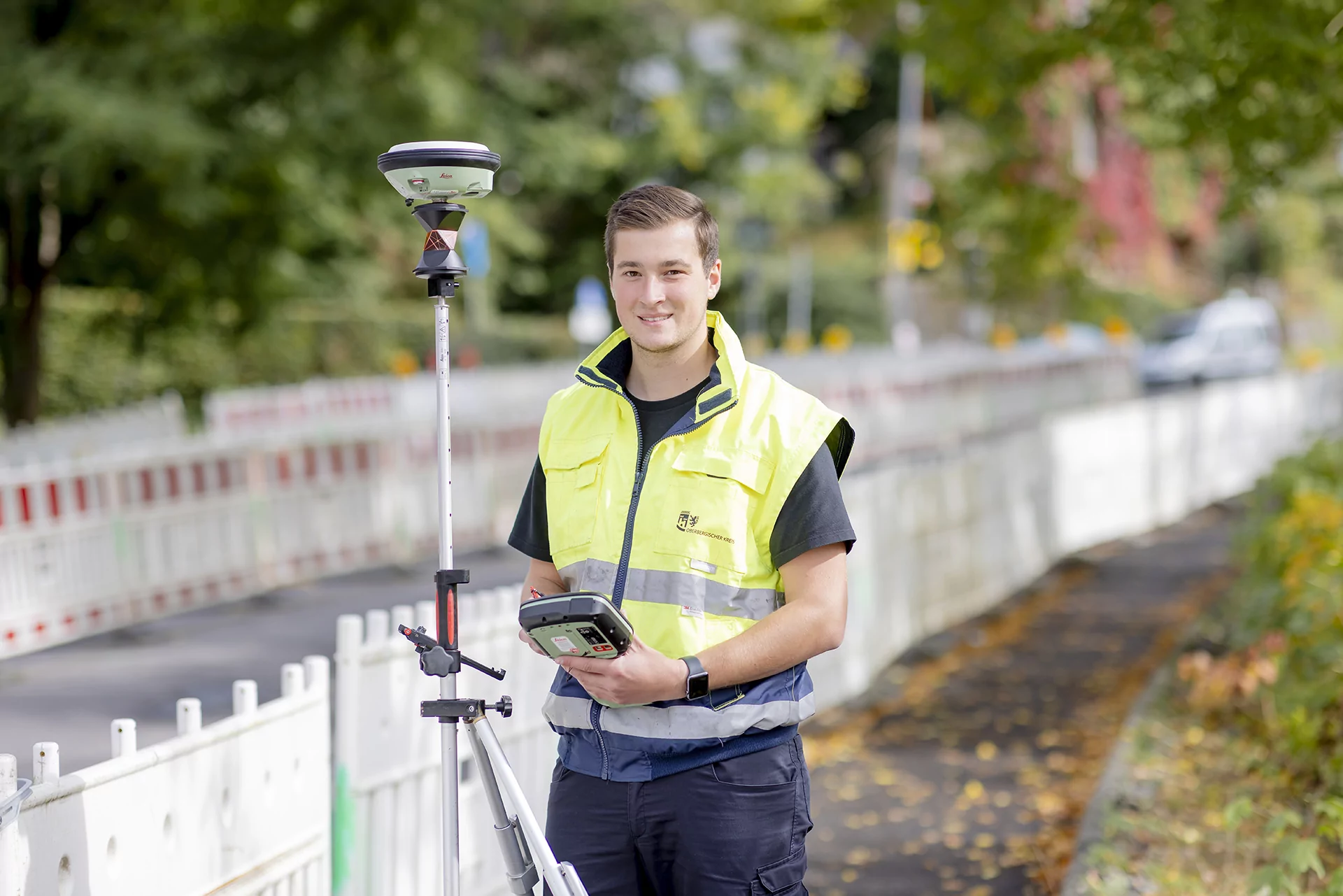 Ein Vermessungstechniker in einer gelben Sicherheitsweste bedient in der N&auml;he einer eingez&auml;unten Stra&szlig;e ein Vermessungsger&auml;t auf einem Stativ.