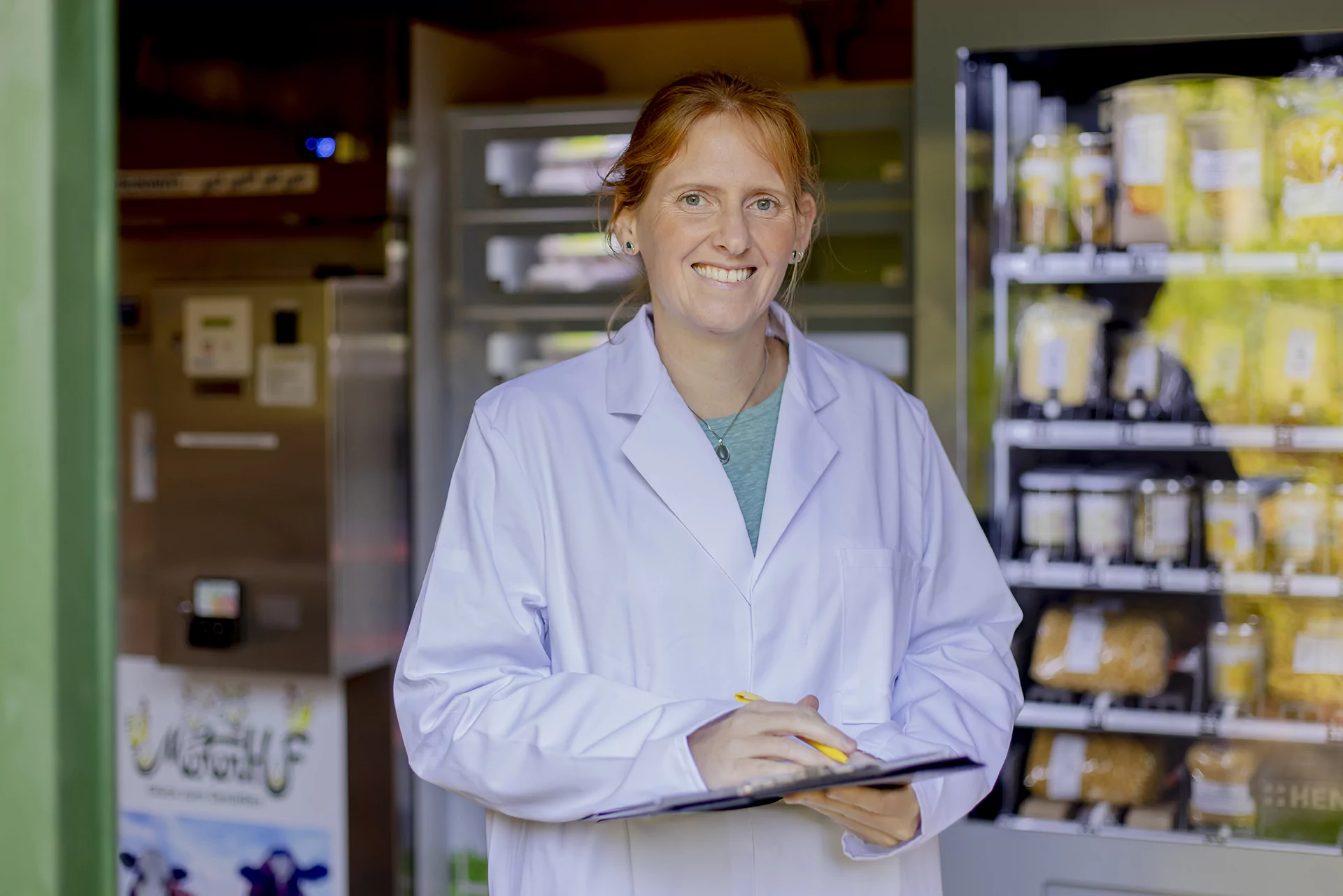 Foto: Katharina Hein Eine Frau steht im weißen Laborkittel und mit Klemmbrett vor einem Verkaufsautomaten mit verschiedenen Lebensmitteln.