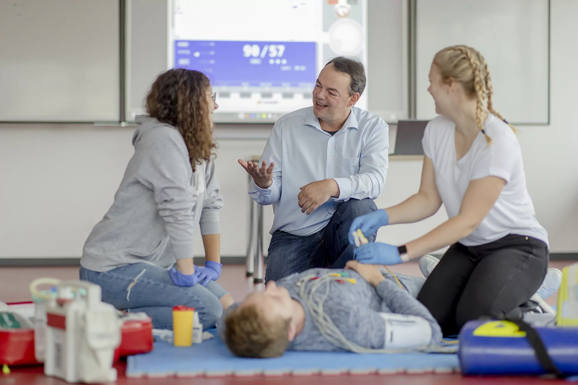 Vier Personen nehmen an einem Erste-Hilfe-Training in einem Schulungsraum teil. Zwei Frauen mit blauen Handschuhen f&uuml;hren eine medizinische &Uuml;bung an einer auf dem Boden liegenden Person durch, w&auml;hrend ein Mann in einem hellblauen Hemd mit einer weiteren Teilnehmerin spricht. Im Hintergrund ist eine Pr&auml;sentation mit medizinischen Werten zu sehen.