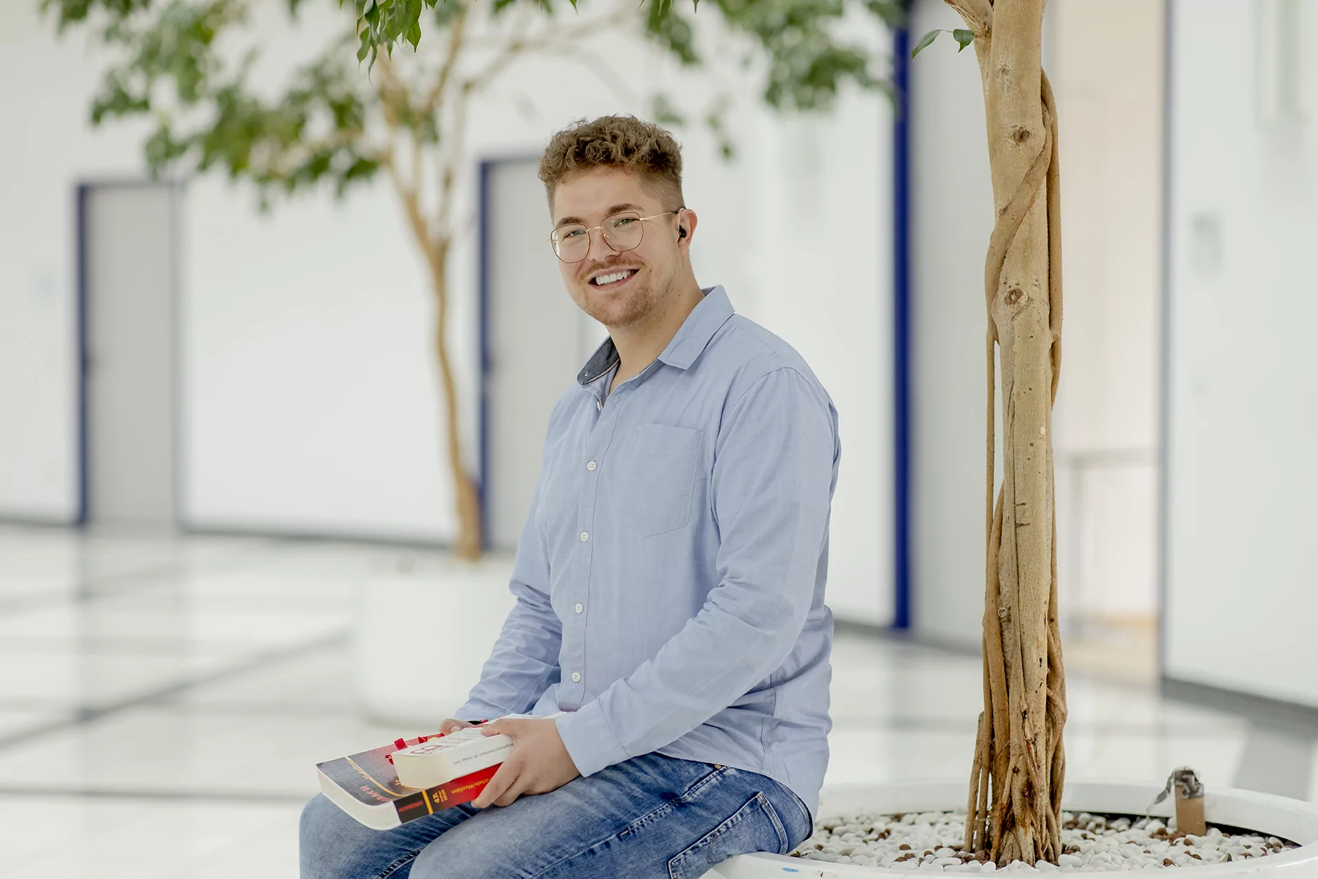 Ein l&auml;chelnder Mann mit Brille und blauem Hemd sitzt in einem Innenraum vor einem Topfbaum und h&auml;lt ein Buch.