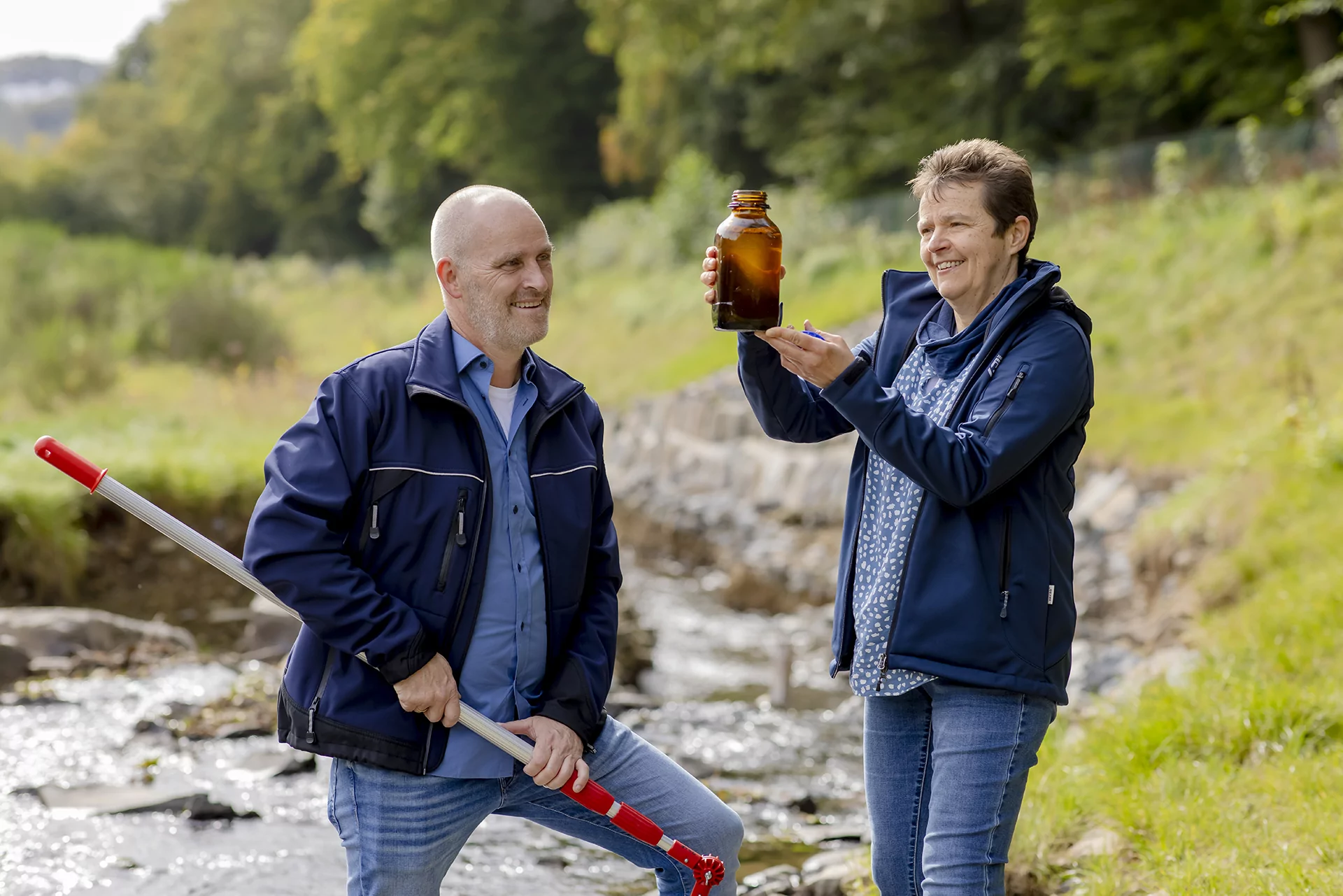 Zwei Menschen in Jacken an einem Bach; einer h&auml;lt eine braune Flasche, der andere eine Stange. Im Hintergrund gr&uuml;ne B&auml;ume.