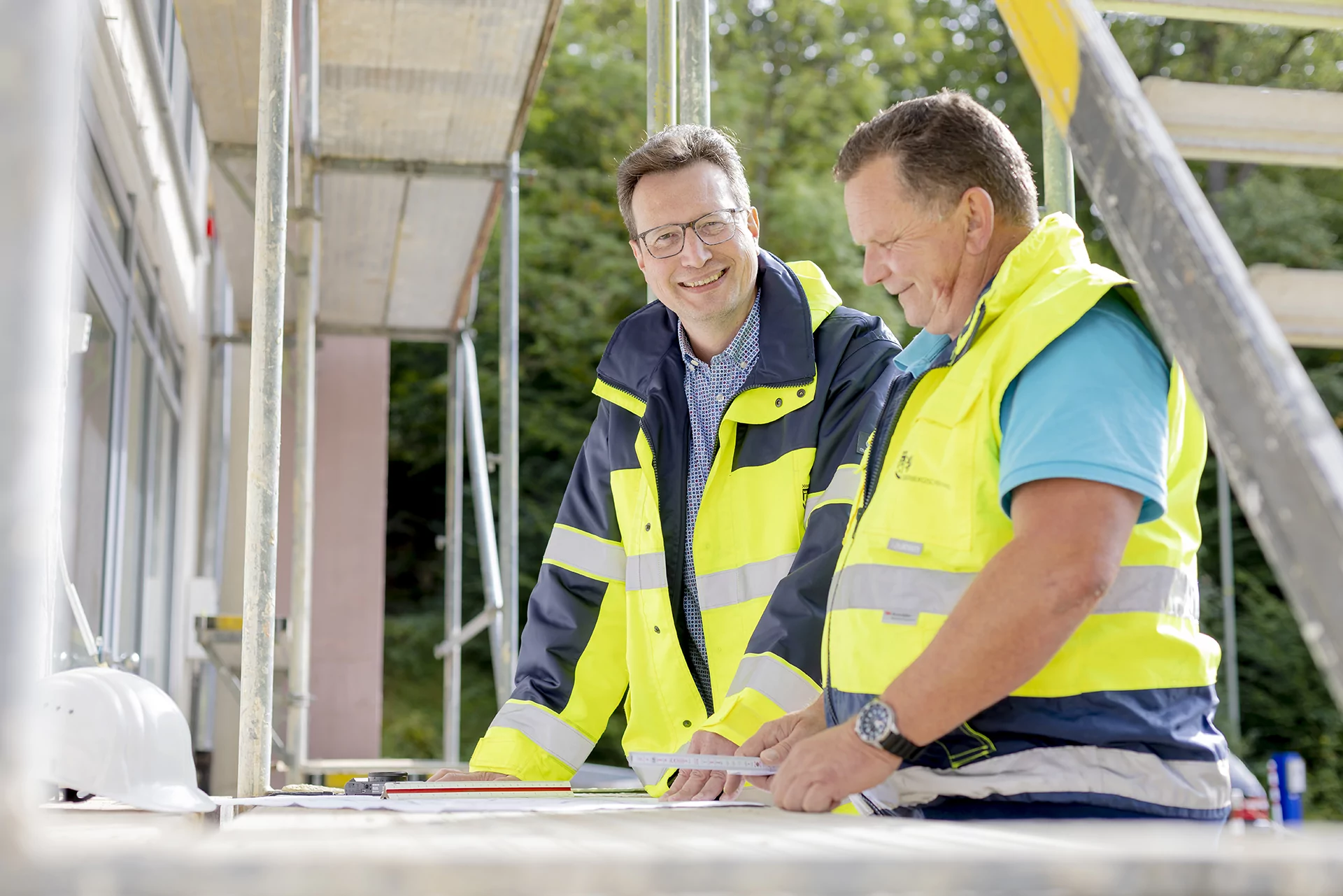 Zwei M&auml;nner in Sicherheitswesten l&auml;cheln und unterhalten sich auf einer Baustelle.