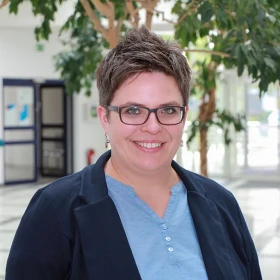 Frau mit kurzen Haaren und Brille, trägt ein blaues Hemd und eine dunkle Jacke und lächelt in einer hellen Innenumgebung mit Pflanzen im Hintergrund.