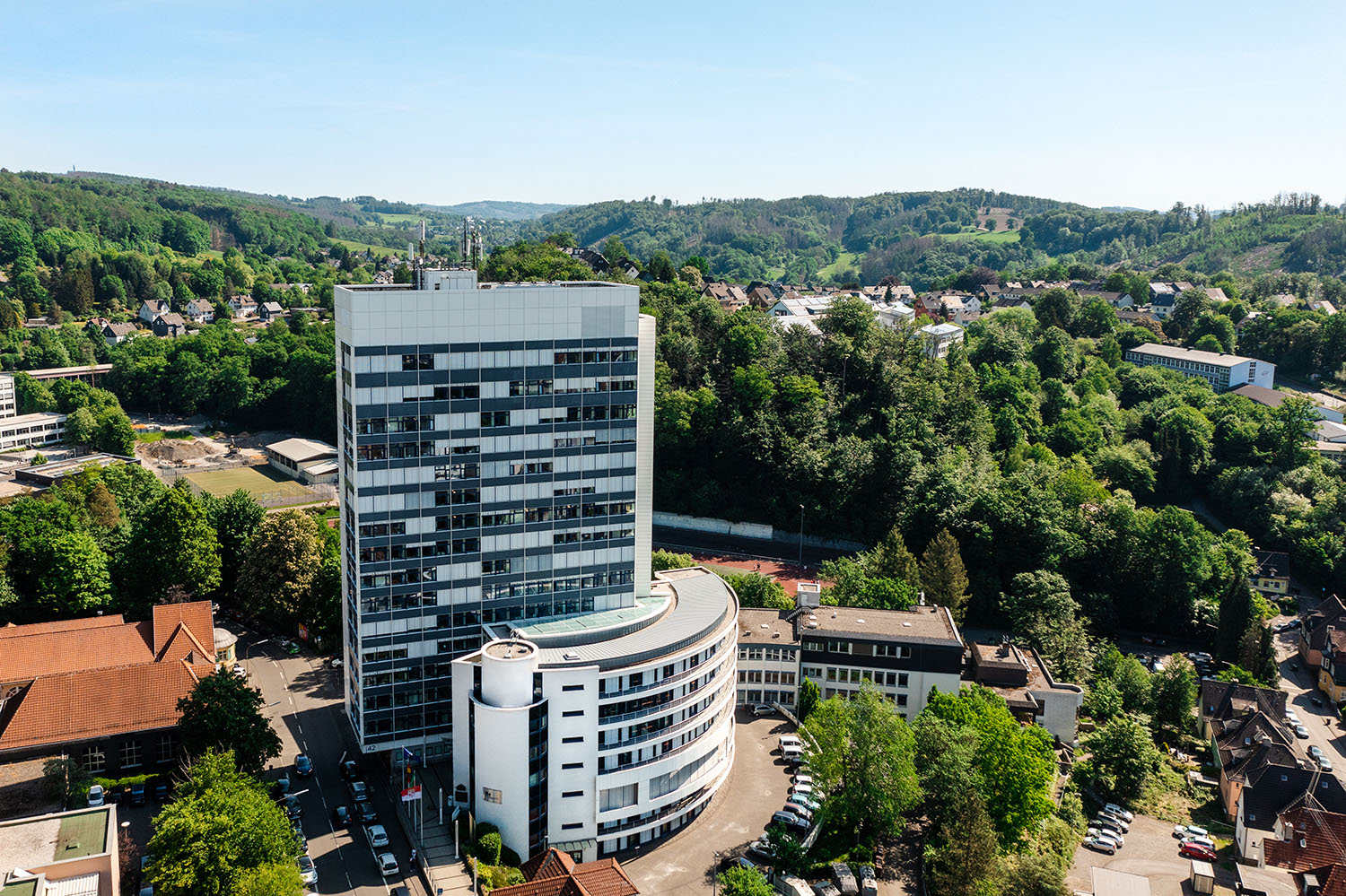 Foto: Jost Gabriel (Dronauten) Das Bild zeigt das Kreishaus in Gummersbach aus der Luftperspektive, ein modernes, mehrstöckiges Gebäude mit rundem Anbau, umgeben von Bäumen, Hügeln und einer Grünanlage unter klarem Himmel.