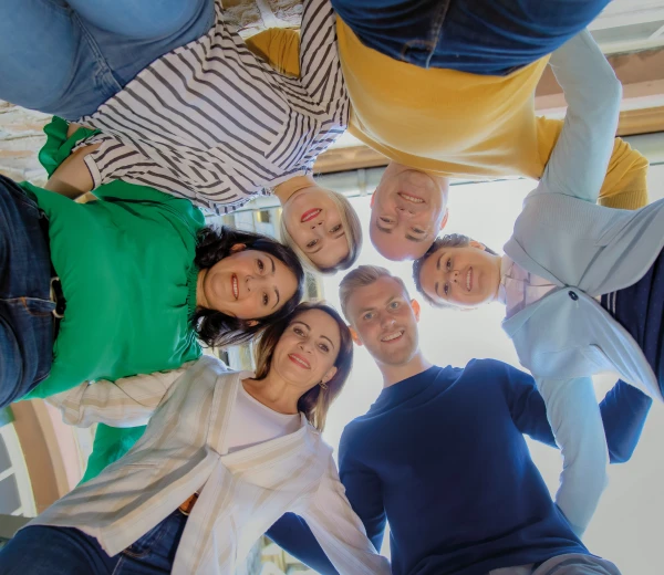 Eine Gruppe von sechs Personen steht draußen in einem Kreis und blickt lächelnd nach unten in die Kamera. Sie tragen farbenfrohe Kleidung.