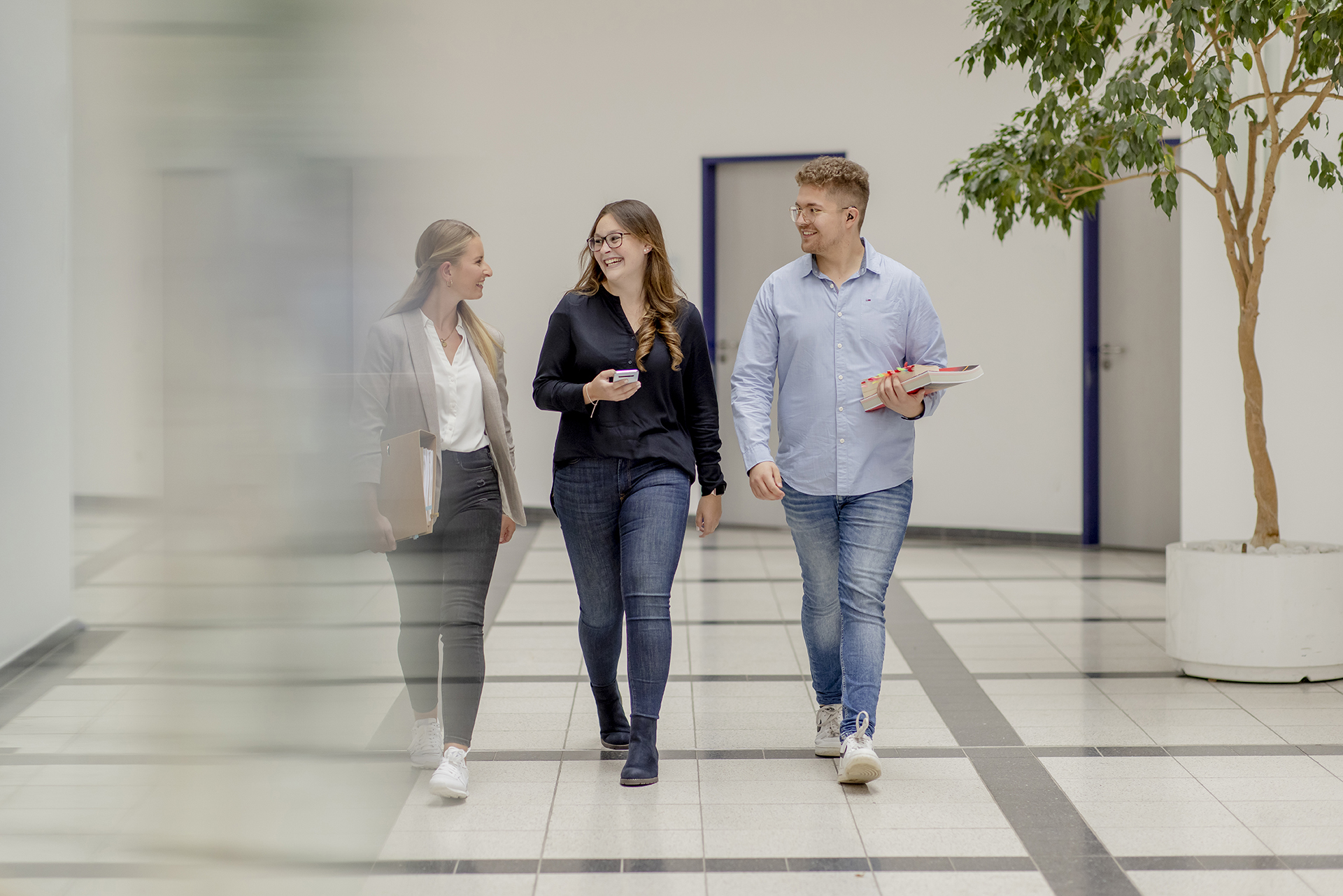 Drei Menschen gehen l&auml;chelnd drinnen umher. Einer h&auml;lt einen Laptop, ein anderer tr&auml;gt B&uuml;cher und der dritte hat ein Telefon. Im Hintergrund Topfb&auml;ume.