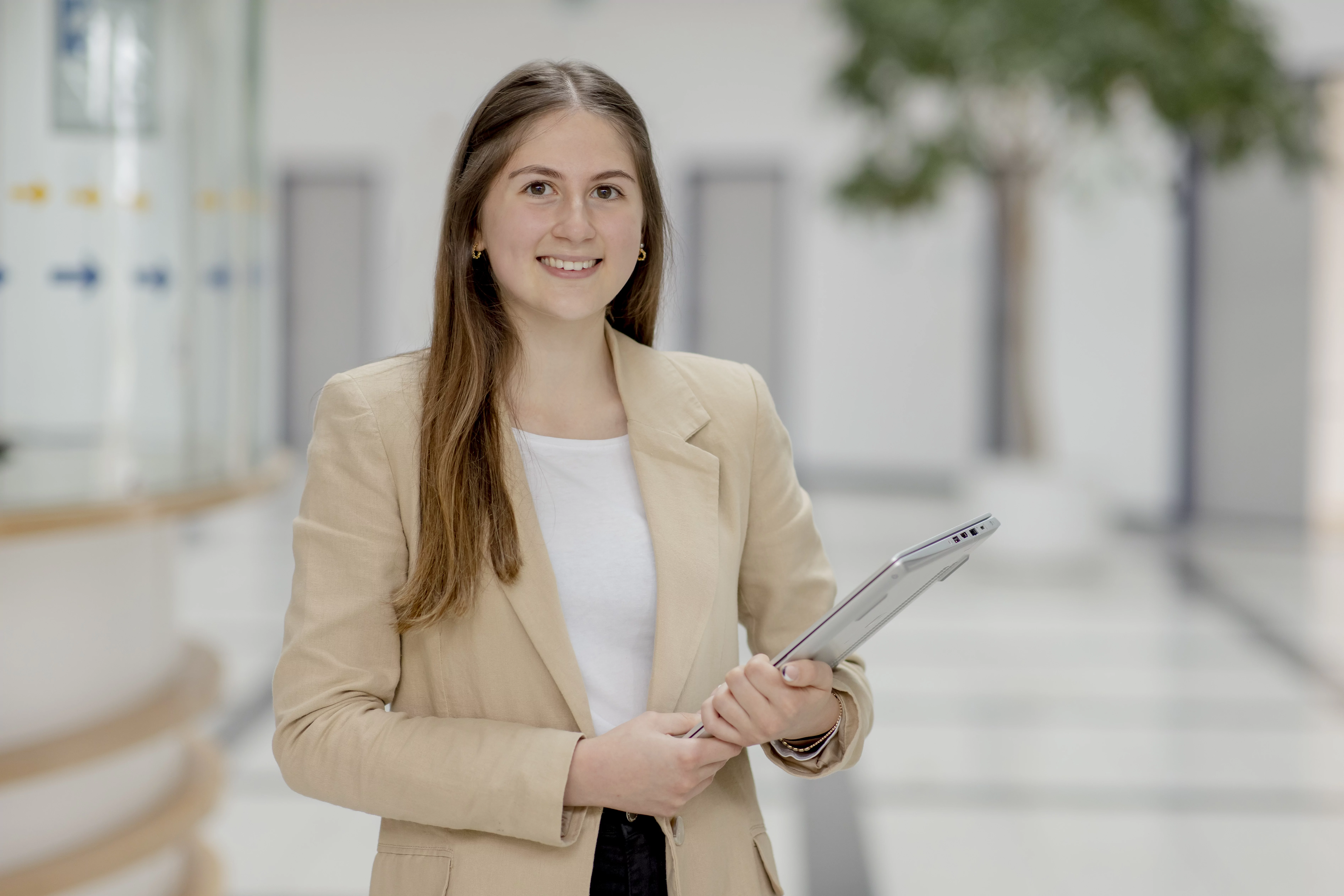Eine l&auml;chelnde Frau in einem beigen Blazer h&auml;lt einige Unterlagen und steht in einem hellen, modernen Flur mit unscharfem Hintergrund.