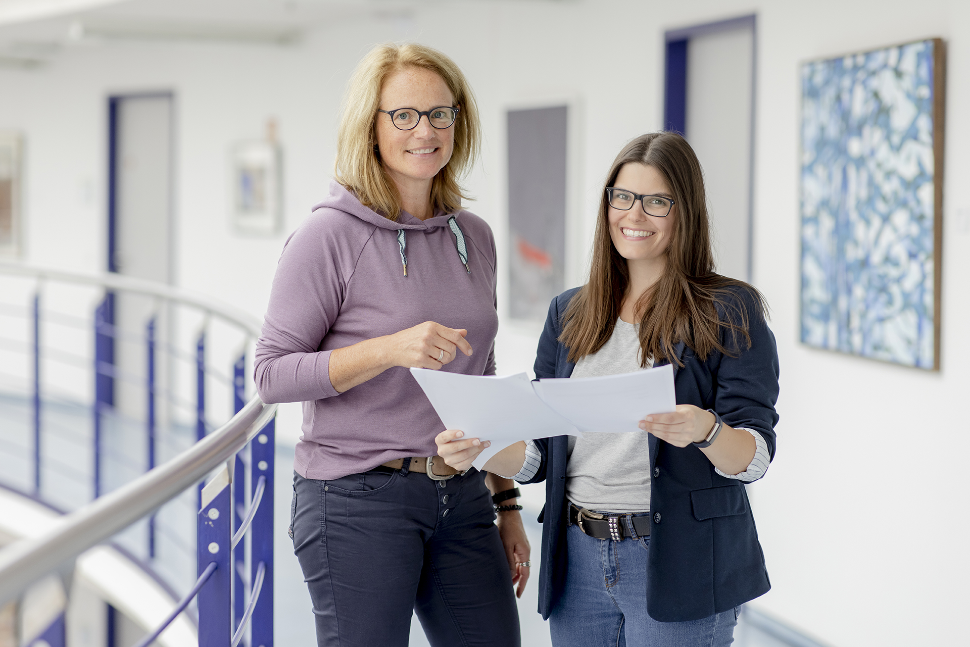 Zwei Frauen stehen in einem modernen B&uuml;roflur, halten Papiere in der Hand und besprechen sie. Sie l&auml;cheln und tragen legere Gesch&auml;ftskleidung.