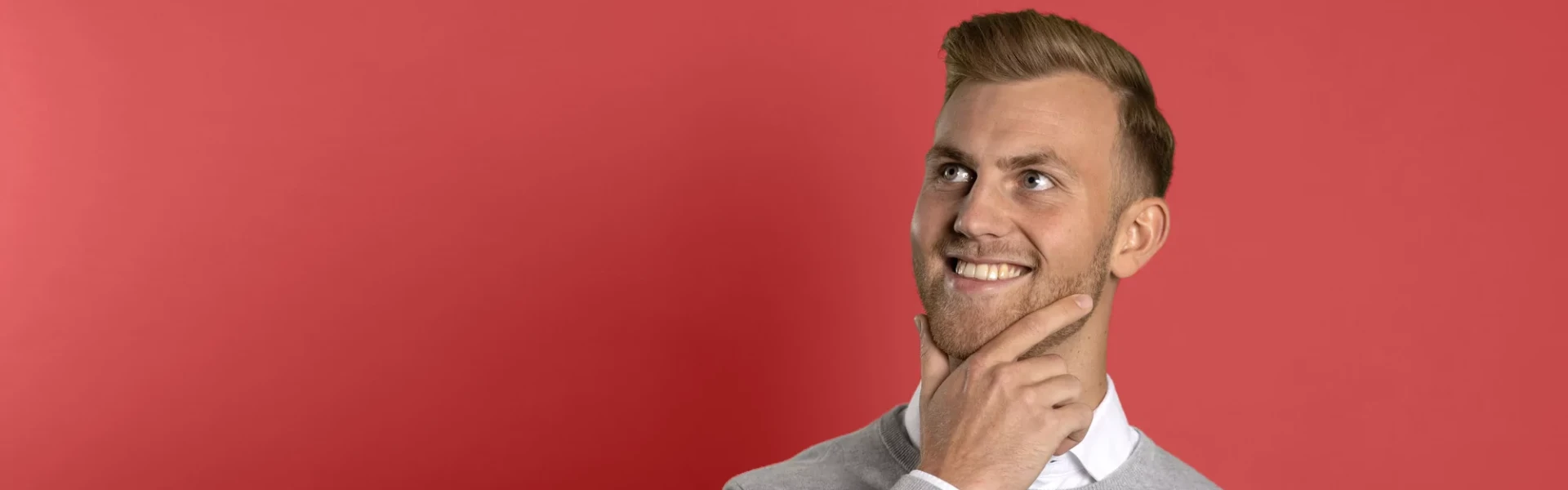 Lächelnde Person in einem grauen Pullover vor einem roten Hintergrund, die nach oben blickt und eine Hand auf dem Kinn hat.