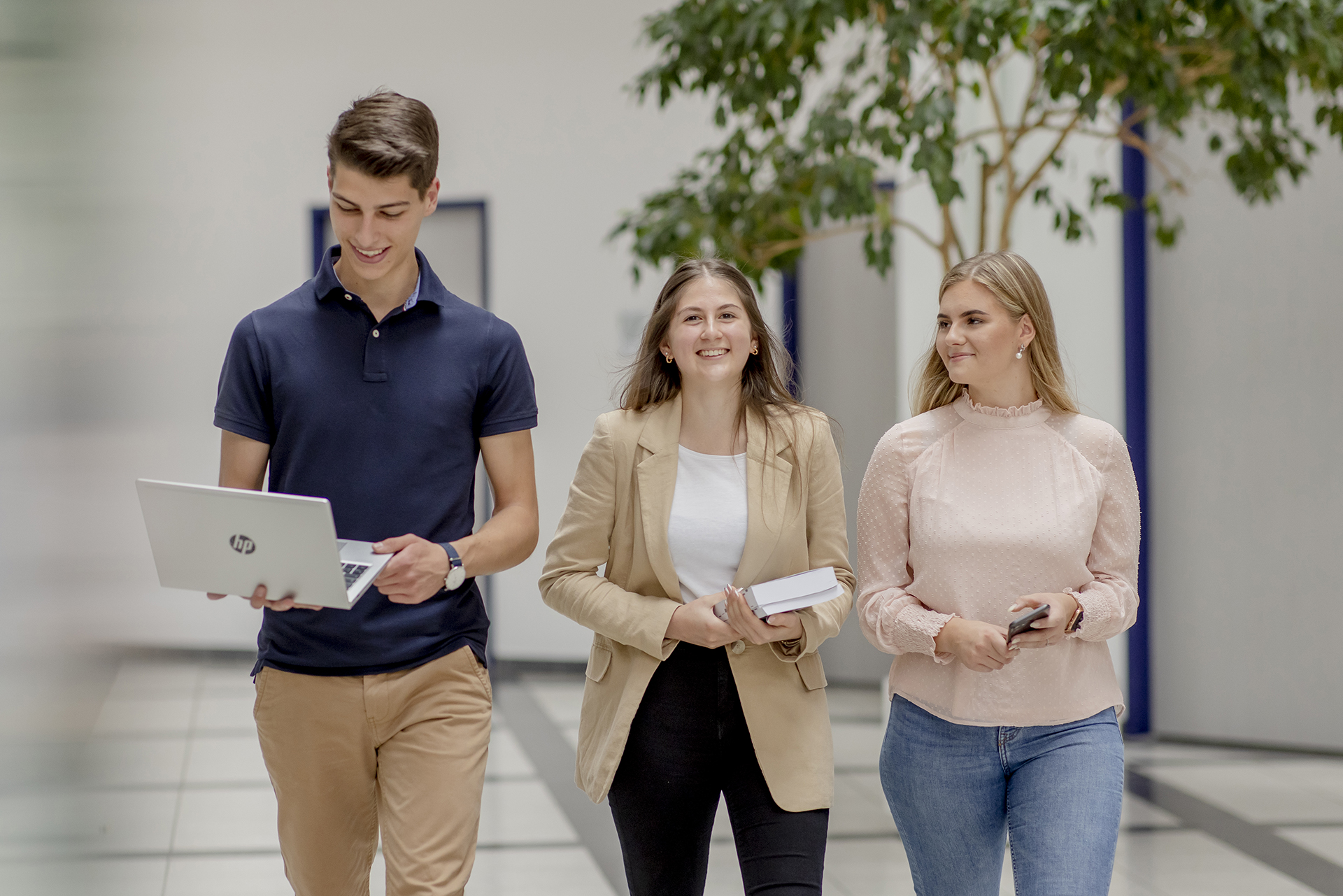 Drei Menschen gehen l&auml;chelnd drinnen umher. Einer h&auml;lt einen Laptop, ein anderer tr&auml;gt B&uuml;cher und der dritte hat ein Telefon. Im Hintergrund Topfb&auml;ume.