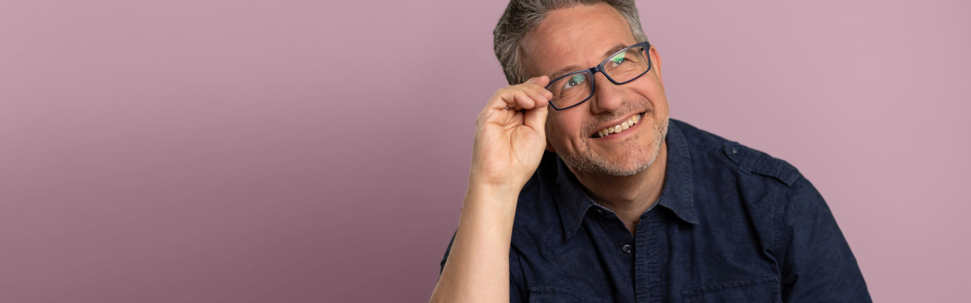 Lächelnder Mann mit Brille sitzt in einem blauen Hemd vor einem rosa Hintergrund und schaut in die Ferne.