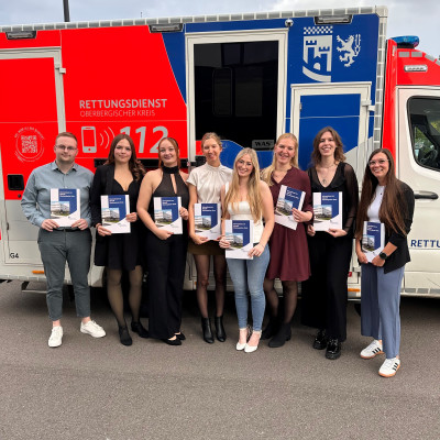 Acht junge Menschen stehen vor einem Rettungswagen und halten ihre Urkunden in die Kamera.