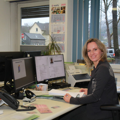 Eine Frau sitzt an einem Schreibtisch mit zwei Bildschirmen. Vor ihr liegen ein paar Unterlagen und sie lächelt freundlich in die Kamera.