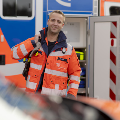 Ein Sanitäter in orange-blauer Uniform steht lächelnd vor einem Krankenwagen und trägt eine Arzttasche.